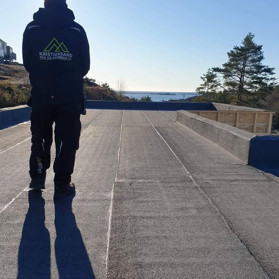 Taktekker i arbeidsklær på et ferdig flatt tak, sjøen i bakgrunnen